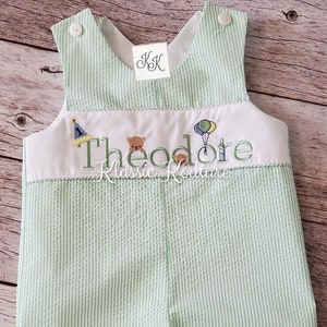 May include: A green and white striped baby overall with a white embroidered bib. The bib features the name "Theodore" with a birthday hat, teddy bear, and balloons.