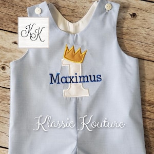 May include: A blue and white gingham baby romper with a white number one and a gold crown embroidered on the front. The name Maximus is embroidered below the number one. The text Klassic Kouture is embroidered on the bottom of the romper.