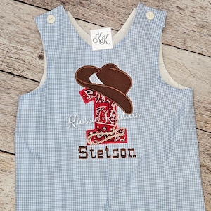 May include: Blue and white gingham baby overalls with a red bandana and brown cowboy hat embroidered on the front. The number "1" is embroidered in the center of the bandana. The name "Stetson" is embroidered below the bandana.