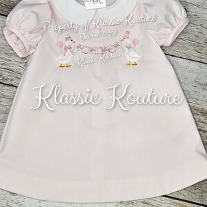 May include: A pale pink baby dress with a white collar and short, puffed sleeves. The dress has an embroidered design of two geese holding balloons and a banner reading "One." The text "Klassic Kouture" is also visible.