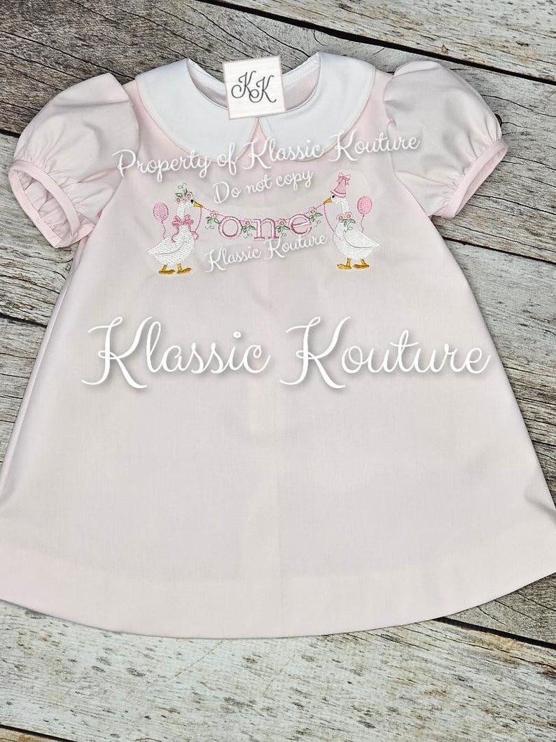 May include: A pale pink baby dress with a white collar and short, puffed sleeves. The dress has an embroidered design of two geese holding balloons and a banner reading "One." The text "Klassic Kouture" is also visible.