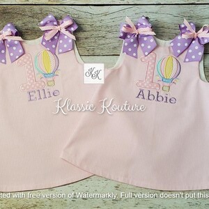 May include: Two pink sleeveless dresses with white polka dots. Each dress has a purple and white bow on the shoulder, a hot air balloon with a number one and the name "Ellie" or "Abbie" embroidered on the front.
