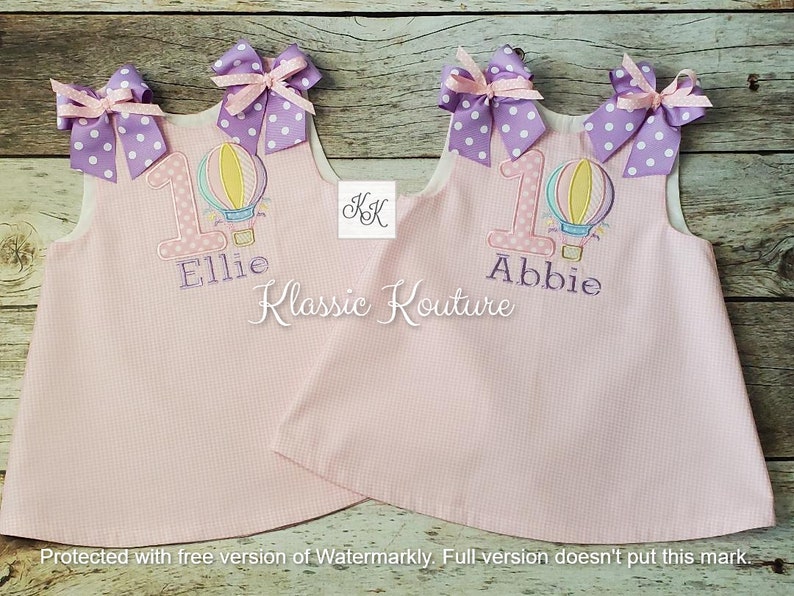 May include: Two pink sleeveless dresses with white polka dots. Each dress has a purple and white bow on the shoulder, a hot air balloon with a number one and the name "Ellie" or "Abbie" embroidered on the front.