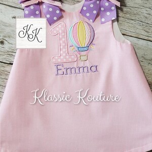 May include: Pink gingham dress with a white hot air balloon and the number 1 embroidered on the front. The dress also has a purple bow with white polka dots on each shoulder. The name "Emma" is embroidered below the number 1. The text "Klassic Kouture" is embroidered on the bottom of the dress.