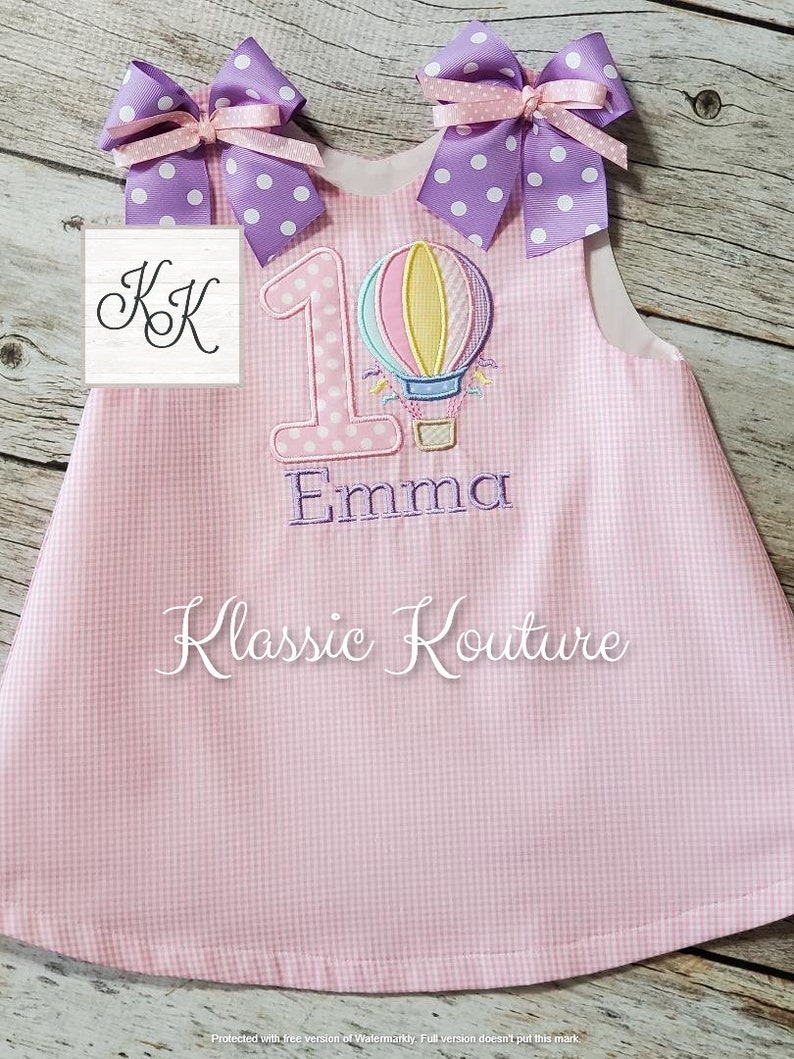May include: Pink gingham dress with a white hot air balloon and the number 1 embroidered on the front. The dress also has a purple bow with white polka dots on each shoulder. The name "Emma" is embroidered below the number 1. The text "Klassic Kouture" is embroidered on the bottom of the dress.