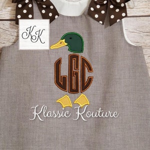 May include: A brown and white gingham sleeveless dress with a brown and white polka dot bow on each shoulder. The dress features an embroidered green duck with a brown monogram "LGC" and the text "Klassic Kouture".