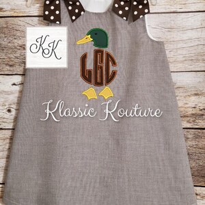 May include: A brown and white gingham dress with a white collar and sleeveless design. The dress features an embroidered duck with a monogram in the centre. The duck is green, brown, and yellow. The dress has two brown and white polka dot bows on the shoulders.