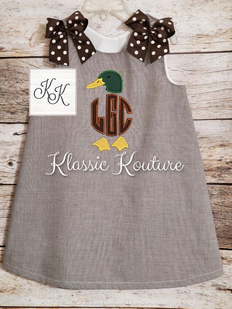 May include: A brown and white gingham dress with a white collar and sleeveless design. The dress features an embroidered duck with a monogram in the centre. The duck is green, brown, and yellow. The dress has two brown and white polka dot bows on the shoulders.