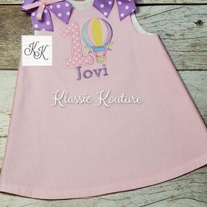 May include: Pink sleeveless dress with a white bodice and a pink and white gingham skirt. The dress has a purple number one with a hot air balloon design and the name "Jovi" embroidered on the front. The dress has two purple bows with white polka dots on the straps.