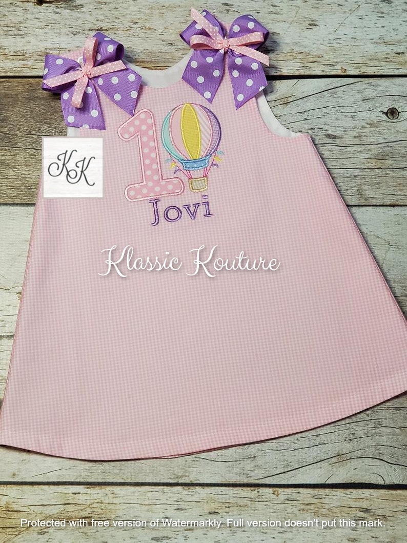 May include: Pink sleeveless dress with a white bodice and a pink and white gingham skirt. The dress has a purple number one with a hot air balloon design and the name "Jovi" embroidered on the front. The dress has two purple bows with white polka dots on the straps.