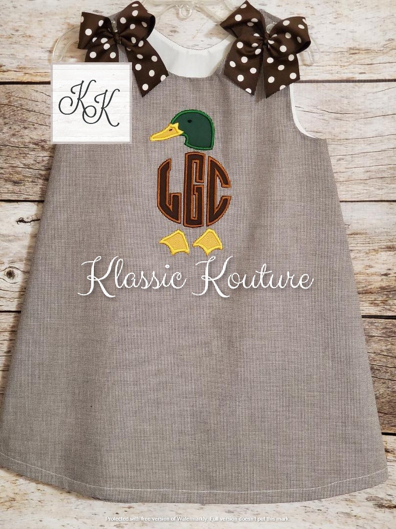 May include: A brown and white gingham dress with a white collar and brown polka dot bows. The dress features an embroidered duck with a monogram in the centre. The text "Klassic Kouture" is embroidered below the duck.