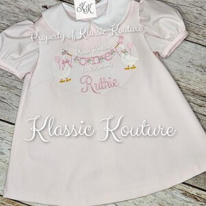 May include: A pale pink baby dress with a white collar and short puff sleeves. The dress has embroidered geese holding balloons and the text "One" and "Ruthie". The brand name "Klassic Kouture" is also visible.