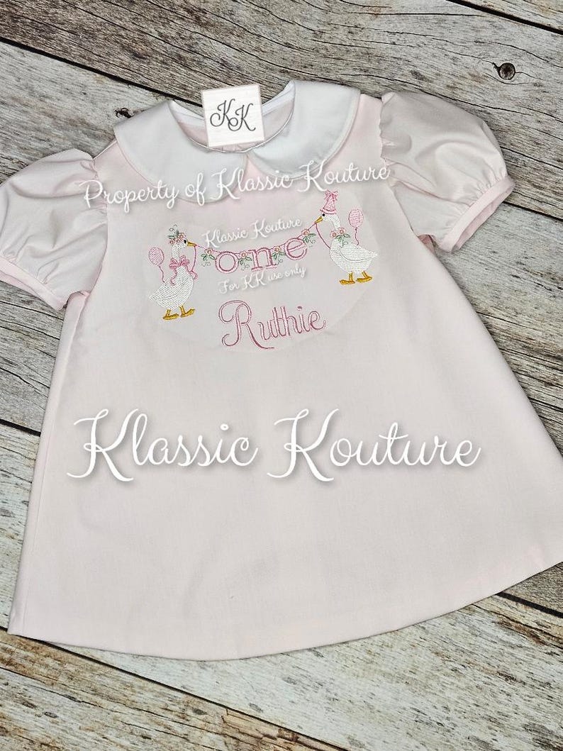 May include: A pale pink baby dress with a white collar and short puff sleeves. The dress has embroidered geese holding balloons and the text "One" and "Ruthie". The brand name "Klassic Kouture" is also visible.