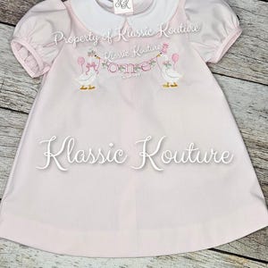 May include: A pale pink baby dress with a white collar and short puff sleeves. The dress has embroidered geese holding balloons and a banner that says "One". The text "Klassic Kouture" is also visible.