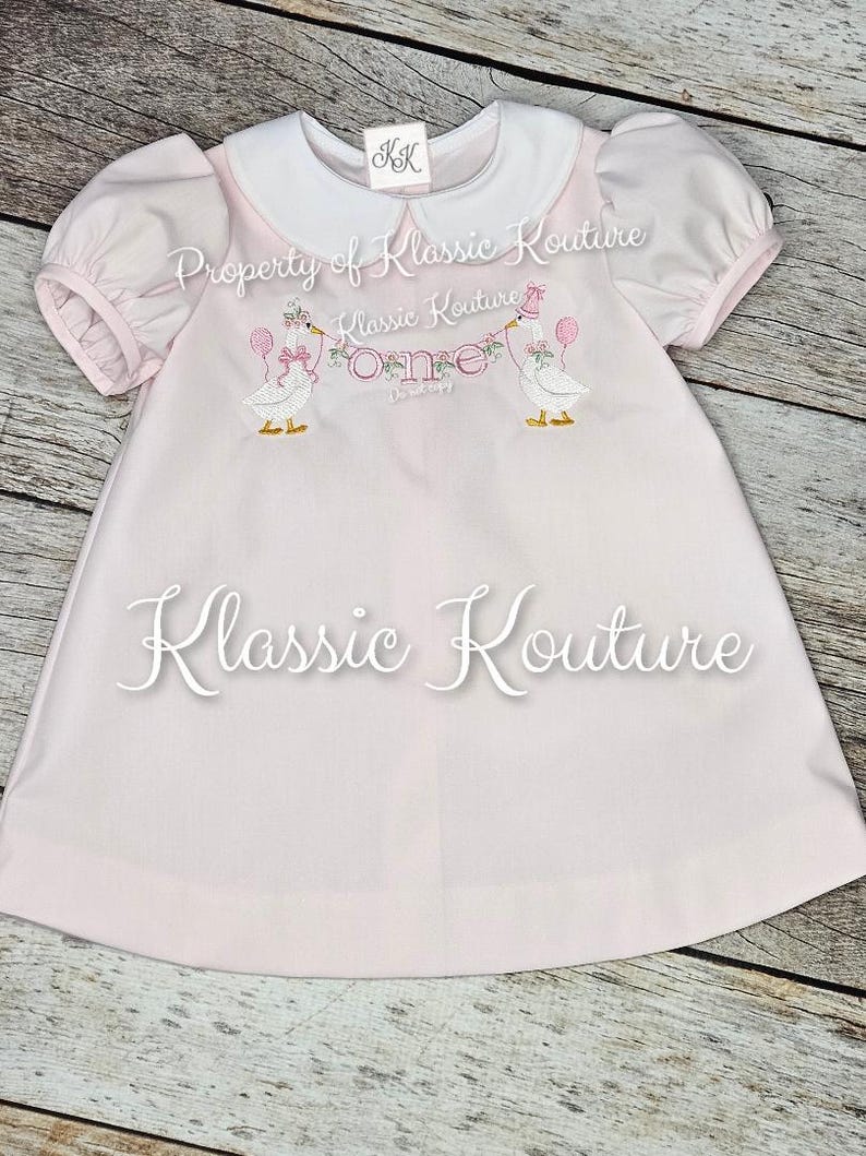 May include: A pale pink baby dress with a white collar and short puff sleeves. The dress has embroidered geese holding balloons and a banner that says "One". The text "Klassic Kouture" is also visible.