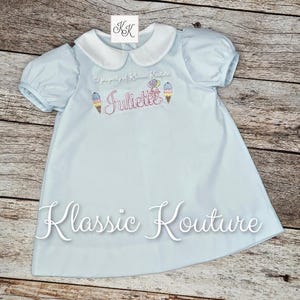 May include: A light blue dress with short puff sleeves and a white Peter Pan collar. The chest features embroidered text reading Juliette, flanked by two ice cream cone designs. The text Klassic Kouture is printed in white across the lower portion.