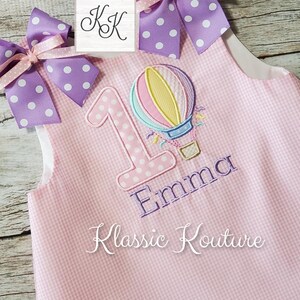 May include: Pink and white gingham fabric with a purple and white polka dot bow. The fabric has a large number "1" with white polka dots, a hot air balloon, and the name "Emma" embroidered on it. The text "Klassic Kouture" is also embroidered on the fabric.