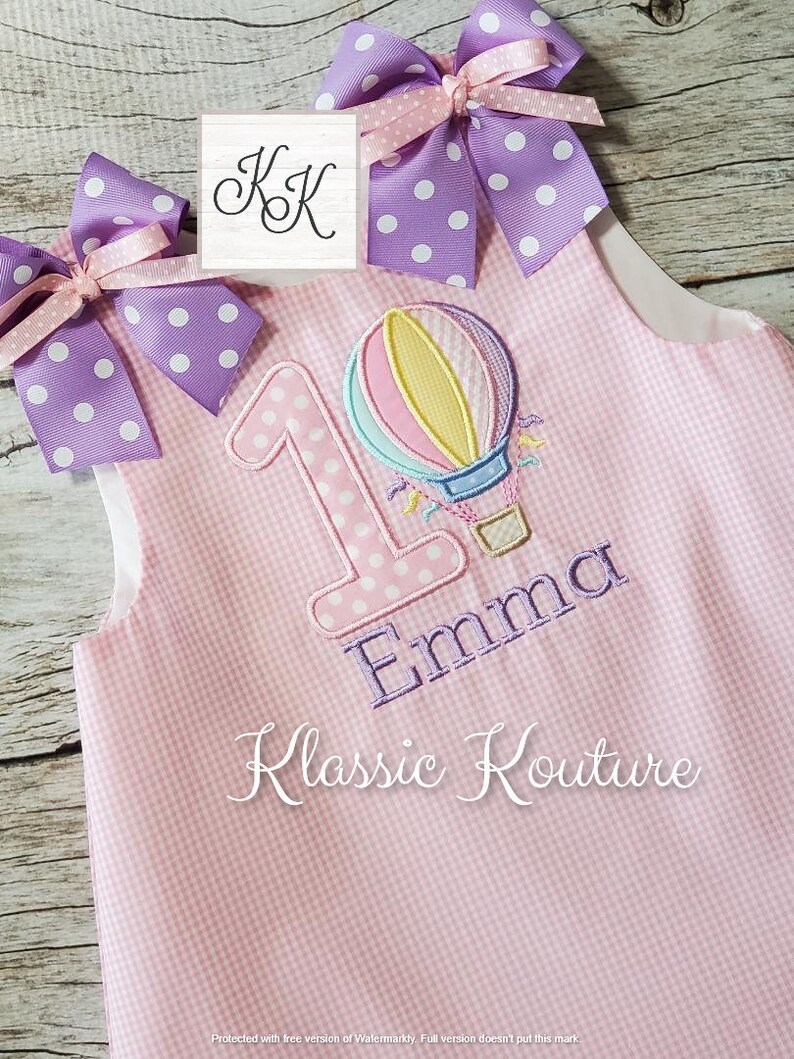 May include: Pink and white gingham fabric with a purple and white polka dot bow. The fabric has a large number "1" with white polka dots, a hot air balloon, and the name "Emma" embroidered on it. The text "Klassic Kouture" is also embroidered on the fabric.