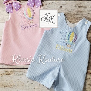 May include: Two personalized baby outfits with a hot air balloon design. The pink outfit has the name "Emma" and the blue outfit has the name "Elliott". Both outfits have a number "1" on them.