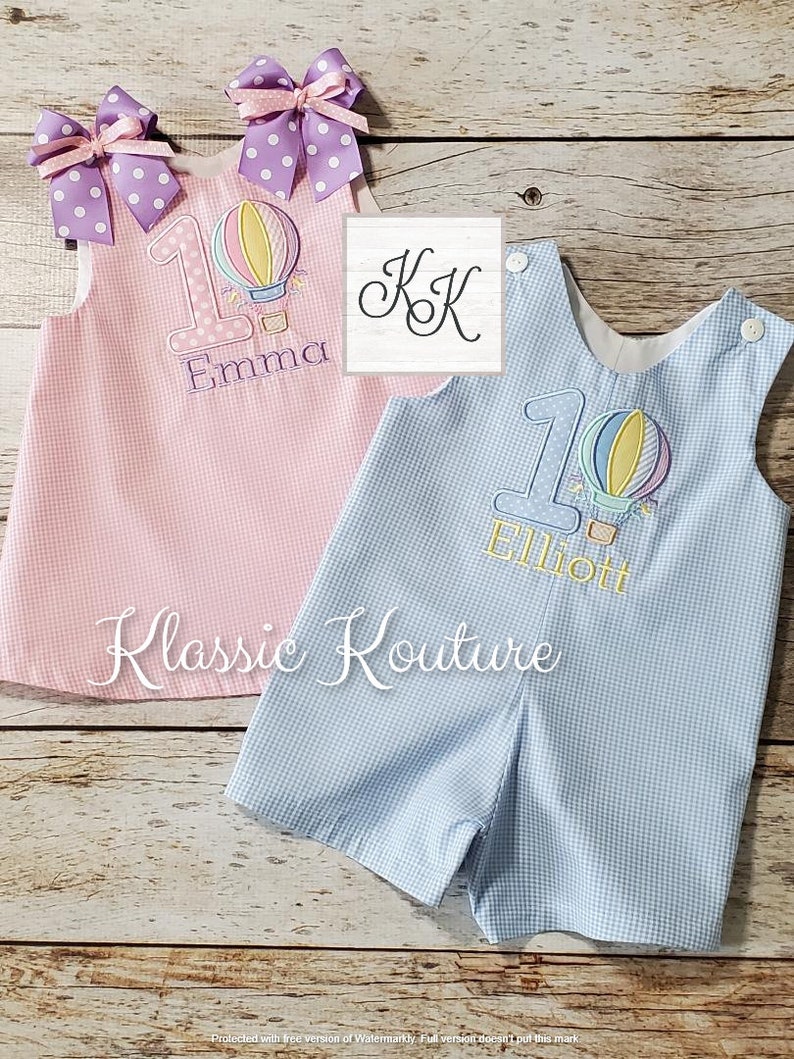 May include: Two personalized baby outfits with a hot air balloon design. The pink outfit has the name "Emma" and the blue outfit has the name "Elliott". Both outfits have a number "1" on them.