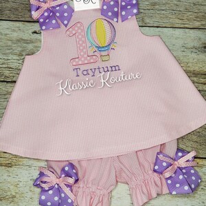 May include: Pink and white gingham baby outfit with a hot air balloon and the number 1 embroidered on the bodice. The outfit also includes a pair of matching bloomers. The text "Taytum Klassic Kouture" is embroidered below the number 1.