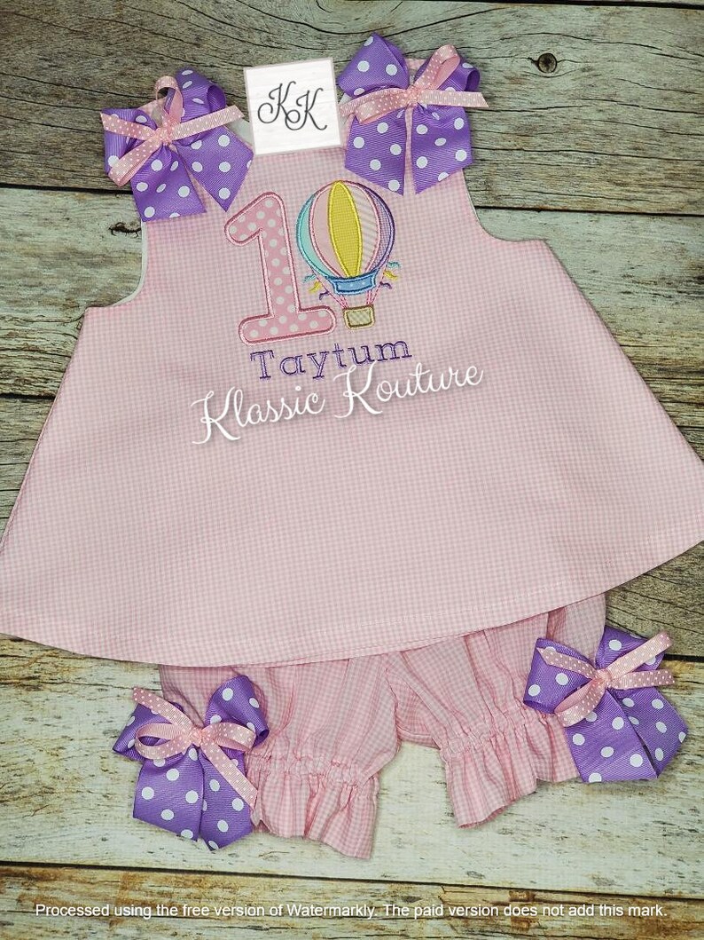 May include: Pink and white gingham baby outfit with a hot air balloon and the number 1 embroidered on the bodice. The outfit also includes a pair of matching bloomers. The text "Taytum Klassic Kouture" is embroidered below the number 1.