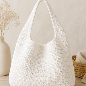 Op de afbeelding: Een witte, gehaakte hobo-tas met een brede, gebogen handgreep. De tas heeft een afgeronde bodem en een getextureerd oppervlak. De achtergrond omvat een vaas met gedroogde bloemen en een rieten mand.