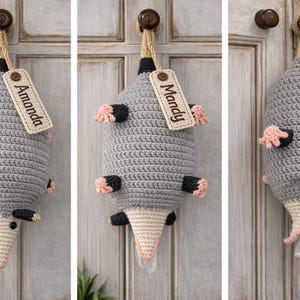 Może przedstawiać: Szydełkowa torba w kształcie oposa z metką z napisem "Amanda". Torba jest szara z czarnymi i różowymi akcentami i wisi na drewnianej gałce od drzwi. Przeznaczona do przechowywania plastikowych toreb.