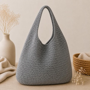 May include: A gray crocheted tote bag with a wide opening and a single handle. The bag has a rounded bottom and a textured surface. The bag is made of a soft, durable material and is suitable for carrying various items.