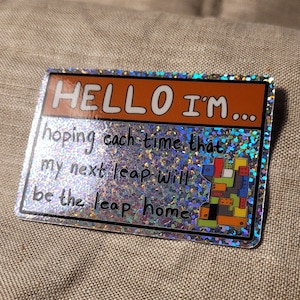 以下が含まれることがあります： 「HELLO I'M... hoping each time that my next leap will be the leap home」というテキストが書かれた銀色のホログラフィックステッカー。ステッカーには、カラフルな積み木が積み重ねられた画像が描かれています。