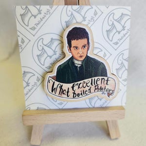 Puede incluir: Recorte de madera de un hombre con abrigo verde con el texto "What excellent boiled Potatoes" sobre un fondo decorativo, presentado en un pequeño caballete de madera.