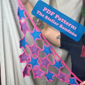 May include: A handmade crochet bandana featuring a star pattern in blue, purple, and pink. The bandana is held up, showcasing the openwork design. Text on a blue label reads "PDF Pattern: The Stellar Bandana."