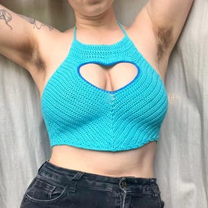 May include: A turquoise crochet halter top with a heart-shaped cutout. The top features a blue trim around the heart and neckline. The top is paired with black denim jeans.