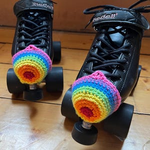 May include: Pair of black roller skates with rainbow-colored crochet toe caps. The skates have black laces and the brand name "Riedell" is visible on the side. The toe caps are in shades of pink, orange, yellow, green, and blue.