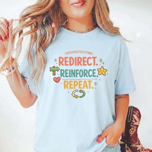May include: Light blue t-shirt with the words "PARAPROFESSIONAL REDIRECT. REINFORCE. REPEAT." in colorful text. The design includes a heart, star, arrow, and recycling symbol. The shirt is a casual style.