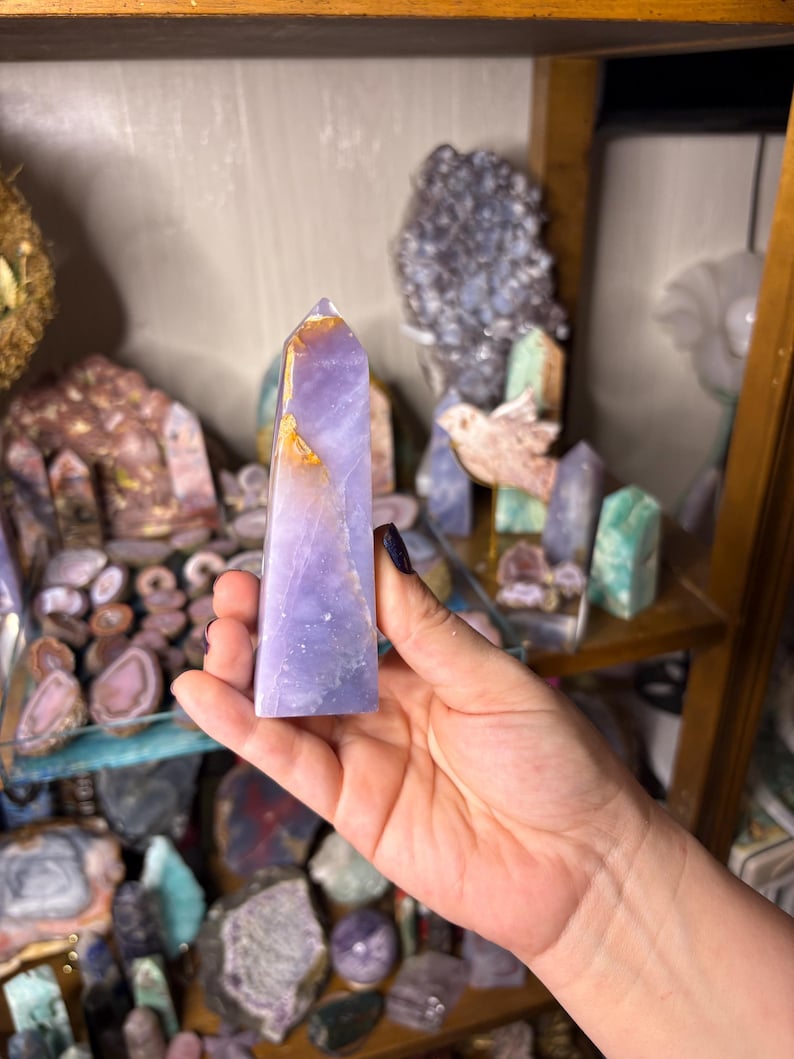 May include: A lavender-colored crystal obelisk with golden and white accents. The crystal is held in a hand, with other crystals and minerals visible on shelves in the background. The obelisk is a polished, pointed shape, and the shelves are made of wood.