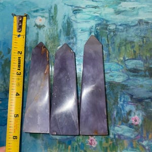 May include: Three purple crystal obelisks, each with a tapered shape and polished surface. The obelisks are approximately 5 inches tall. The crystals have a gradient of color, ranging from light lavender to deep violet.