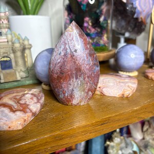 May include: A teardrop-shaped, polished stone with a mix of red, white, and brown hues. The stone is displayed on a wooden shelf alongside other decorative stones and objects. The stone's surface has a natural, marbled appearance.