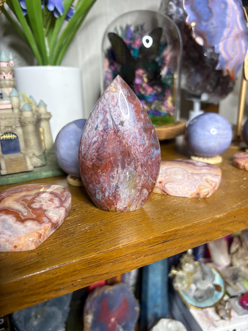 May include: A teardrop-shaped, polished stone with a mix of red, white, and brown hues. The stone is displayed on a wooden shelf alongside other decorative stones and objects. The stone's surface has a natural, marbled appearance.
