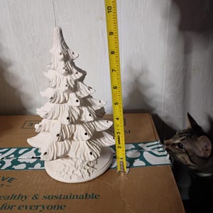 Könnte beinhalten: Ein weißer Weihnachtsbaum aus Keramik, etwa 20 cm hoch, mit strukturierter Oberfläche. Der Baum hat mehrere Äste mit kleinen Löchern für Lichter. Ein gelbes Maßband befindet sich neben dem Baum.