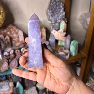 May include: A light purple crystal tower with a pointed top is held in a hand. The crystal has a marbled appearance with lighter and darker shades of purple. Other crystals and minerals are visible on shelves in the background.