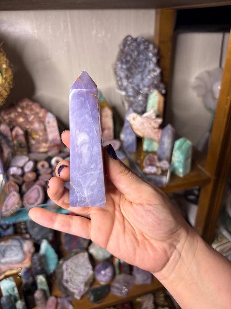 May include: A light purple crystal tower with a pointed top is held in a hand. The crystal has a marbled appearance with lighter and darker shades of purple. Other crystals and minerals are visible on shelves in the background.