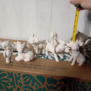 May include: A collection of unpainted ceramic figurines, including a deer, goat, and a figure riding a reindeer. The figurines are arranged on a cardboard box. A measuring tape shows the height of one of the figurines is approximately 4 inches.