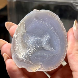 May include: A polished agate slice featuring a translucent light blue and lavender center with concentric banding and a central cluster of sparkling white druzy crystals. The stone has a smooth, rounded edge and a natural, semi-circular shape.