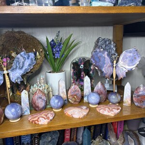 May include: A wooden shelf displays a collection of crystals and decorative items. The shelf holds various polished stones, including spheres, obelisks, and heart-shaped pieces. Two butterfly sculptures with blue and purple wings are also present, along with a small potted plant.
