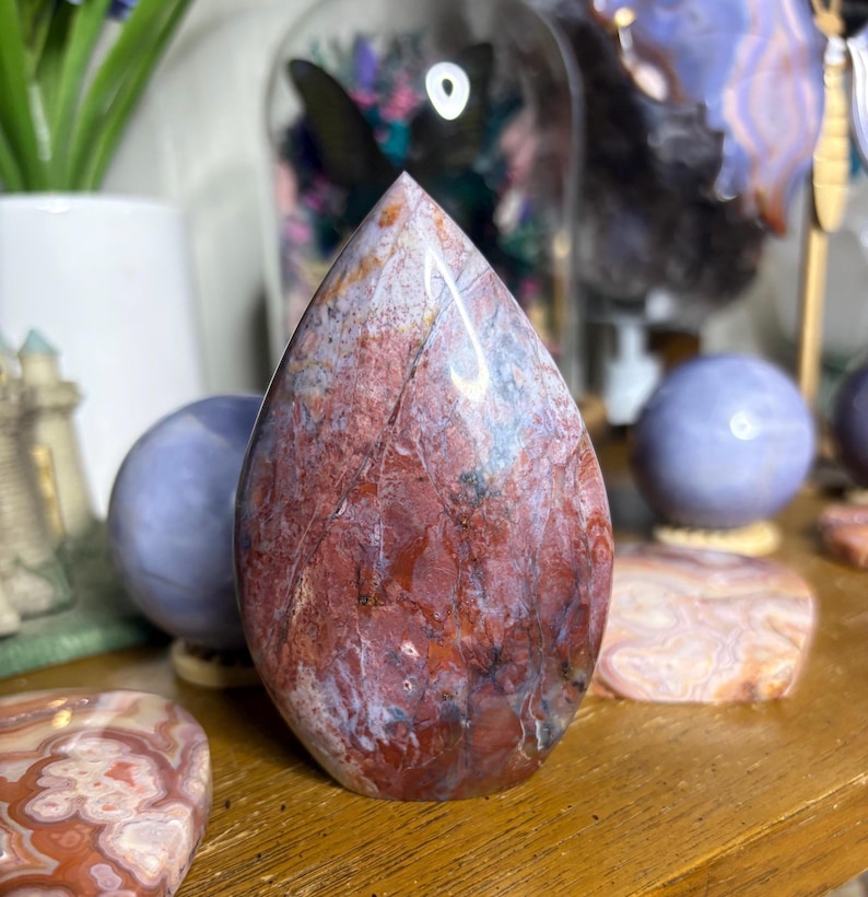 May include: A teardrop-shaped, polished stone with a mix of red, white, and gray hues. The stone has a smooth, glossy surface and a pointed top. Other polished stones and decorative items are in the background.