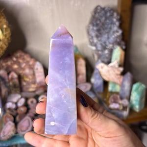 May include: A light purple crystal obelisk with a pointed top. The crystal has a smooth, polished surface with natural variations in color and texture. The obelisk is held in a hand, with other crystals in the background.