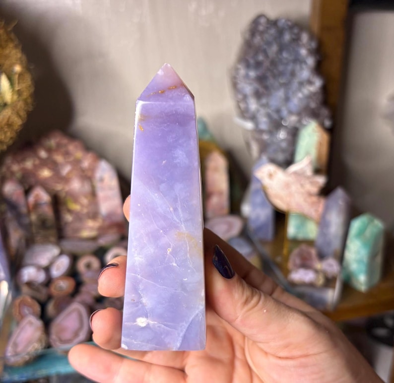 May include: A light purple crystal obelisk with a pointed top. The crystal has a smooth, polished surface with natural variations in color and texture. The obelisk is held in a hand, with other crystals in the background.