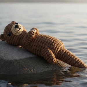 May include: A crocheted brown bear toy rests on a rock in the water. The bear has a light tan muzzle and a black nose and eyes. The bear is lying on its back with its arms crossed, appearing relaxed. The background shows a body of water and mountains.