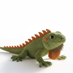 Peut inclure: Un iguane au crochet fait à la main dans les tons de vert, d'orange et de beige. L'iguane a un corps texturé, une rangée d'épines orange sur le dos et un fanon beige. L'iguane est posé sur un fond blanc.